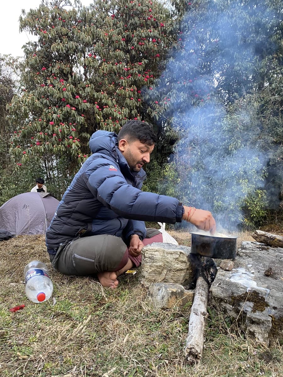 Govinda Subedi cooking over a campfire under rhododendron trees on trek
